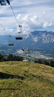A ski lift ascends a snowy mountain in Arnoldstein Dreiländereck, a ski resort in Austria. Surrounding the lift is a challet and a vibrant winter sports scene.