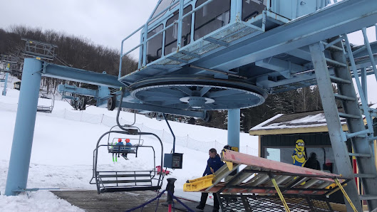 A winter scene at Holimont New York featuring a ski lift carrying skiers up the snowy mountainside at the ski resort. A skier navigates the slope and a visible snowmobile adds excitement.