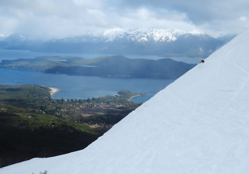 A skier and a snowboarder enjoying a winter sports scene in Cerro Bayo Neuquén Argentina. They're gliding down a snow-covered mountain with a small chalet visible in the distance.