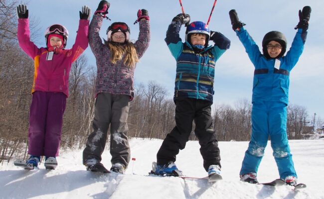 A winter sports scene at Camp Fortune in Quebec, Canada. A family is skiing down snow-covered slopes amidst pine trees, with a ski lift visible in the background.