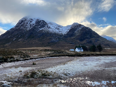 View of a cozy lodge and mountain hut nestled amidst the majestic scenery of Glencoe Mountain in Scotland, which appears to be cloaked in a beautifully serene winter landscape.