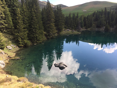 View of the serene lake at Elsigen-Metsch in Frutigen Achseten under a clear sunny day. A charming chalet nestled near the stunning mountain in the Bernese Oberland completes the picture.