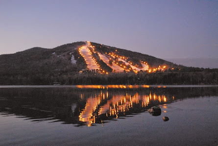 View of Pleasant Mountain in Bridgton, Maine, showcasing a ski resort with a challet. Winter sports enthusiasts can be seen enjoying the snowy slopes against the backdrop of a serene lake.