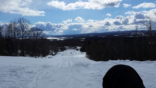 A winter sports scene at Bigrock Mountain in Maine USA showing a skier tackling the snowy slopes of the mountain. A chalet and snowmobile are also visible highlighting the area's recreational activities.