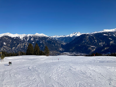 A winter sports scene at Skilifte und Sesselbahn Sarn Heinzenberg in Switzerland. The image includes a skier near a chalet with the ski resort visible in the background.