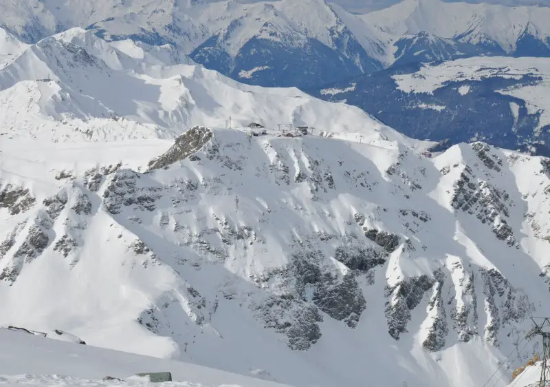 A charming chalet nested at La Plagne ski resort in Savoie Mont Blanc, France. A solitary skier navigates the pristine white slopes under the watchful peaks of the surrounding mountains.