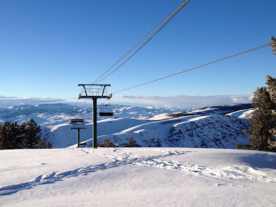 A picturesque winter scene at Pine Creek Wyoming. A ski lift ascends over pristine snowy slopes leading to a chalet nestled amidst snow-covered trees. Ski trails crisscross the landscape near a bustling ski resort.