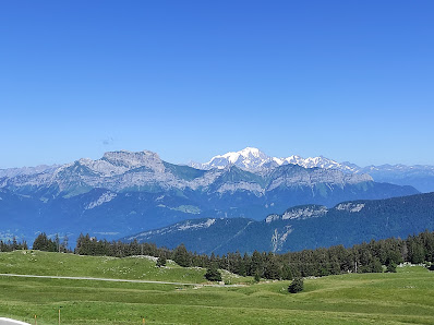 View of the Semnoz mountain station in Savoie Mont Blanc, France. Features a chalet and a ski resort, overlooked by a towering mountain under a clear, sunny sky. A mountain bike is also visible.