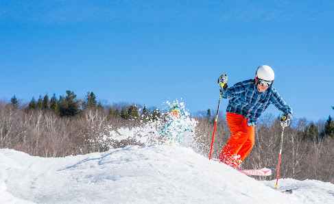 Winter sports enthusiasts enjoying a day on the slopes at Sommet Olympia in Piedmont, Quebec, including an active skier gliding downhill and a snowboarder. Surrounding mountainous landscape of the ski resort is blanketed by fresh snow.